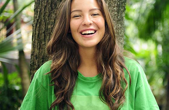 Uma mulher de cabelos castanhos e encaracolados, sorri e usa uma camiseta verde.